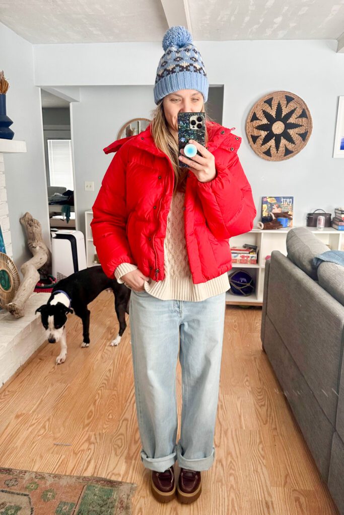Casual February outfit with jeans, a cozy sweater, bright red puffer jacket, knit hat, and boots in snowy weather