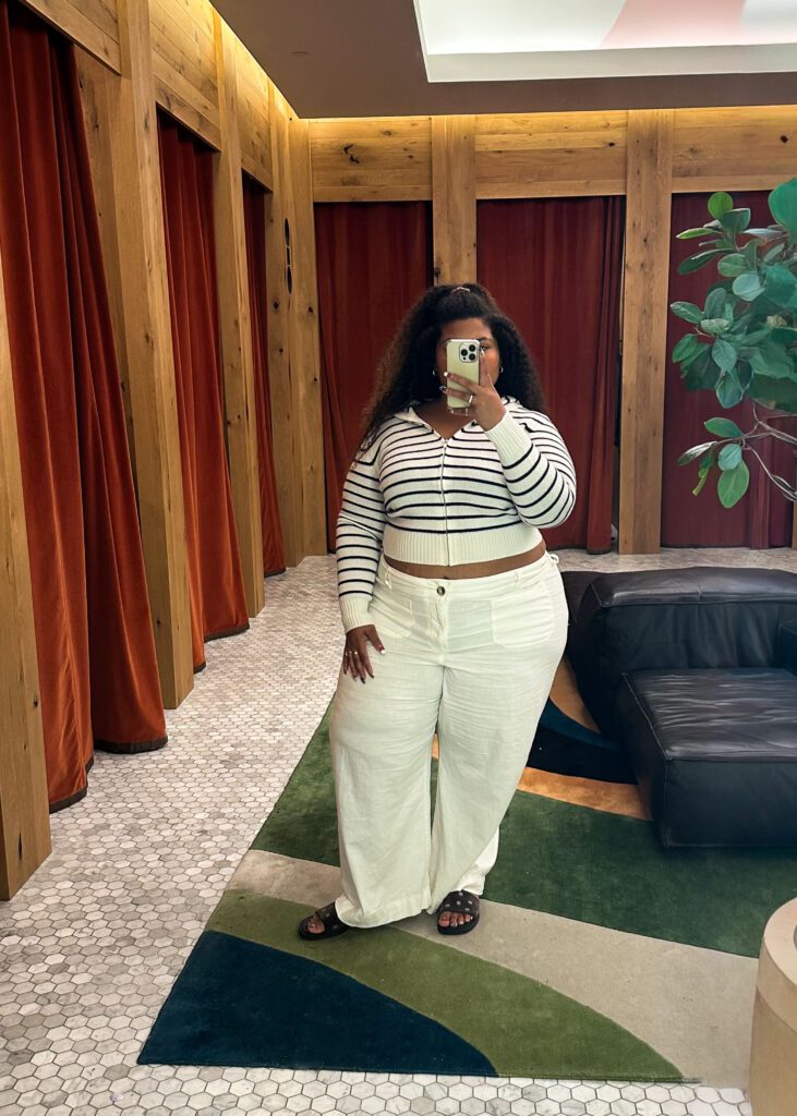 Full-length dressing room selfie of a plus-size woman wearing a striped zip-up cardigan, brown belt, and white jeans at Aritzia.