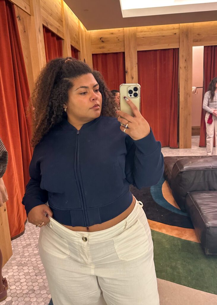 Full-length dressing room selfie of a plus-size woman wearing Aritzia's navy boyfriend bomber zip-up with white jeans.